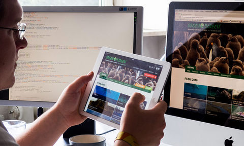 Ein junger Mann schaut sich eine Website auf einem Tablet an. Im Hintergrund ist die selber Website auf einem Desktop-Rechner zu sehen.